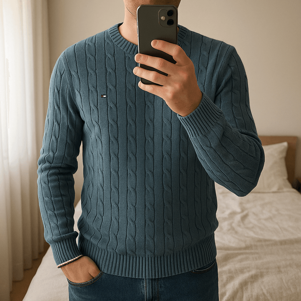 Pull Col Rond Torsadé 100 % Coton Tommy Hilfiger Bleu M debout, face à la caméra, dans une chambre à la lumière douce, avec une lumière naturelle filtrant par la fenêtre.