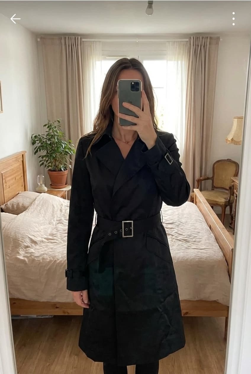 Trench coat Etam debout, face à la caméra, dans une chambre à la lumière douce, avec une lumière naturelle filtrant par la fenêtre.