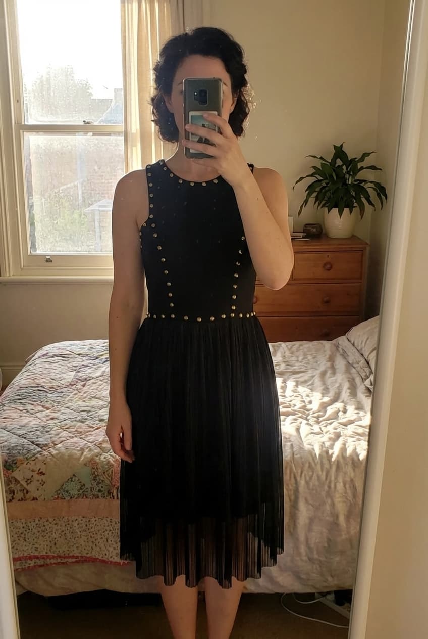 Joloe robe noire debout, face à la caméra, dans une chambre à la lumière douce, avec une lumière naturelle filtrant par la fenêtre.