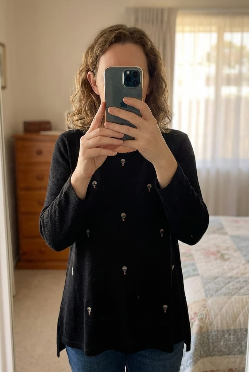 Haut / blouse noir avec broderies TXl debout, face à la caméra, dans une chambre à la lumière douce, avec une lumière naturelle filtrant par la fenêtre.