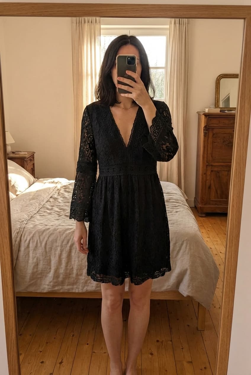 Robe dentelle noir S debout, face à la caméra, dans une chambre à la lumière douce, avec une lumière naturelle filtrant par la fenêtre.