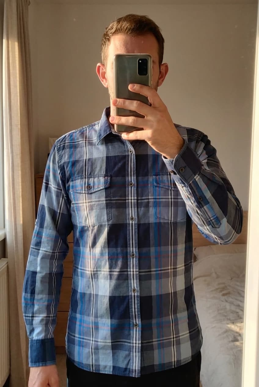 Une personne en Chemise manche longue Taille L debout, face à la caméra, dans une chambre à la lumière douce, avec une lumière naturelle filtrant par la fenêtre.