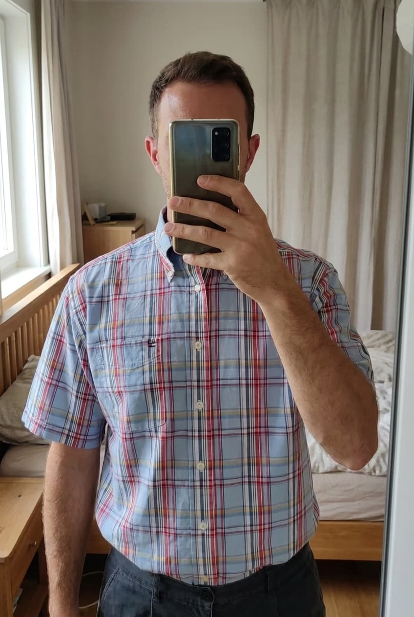 Une personne en Chemise manche courte, tommy Hilfiger, taille L debout, face à la caméra, dans une chambre à la lumière douce, avec une lumière naturelle filtrant par la fenêtre.