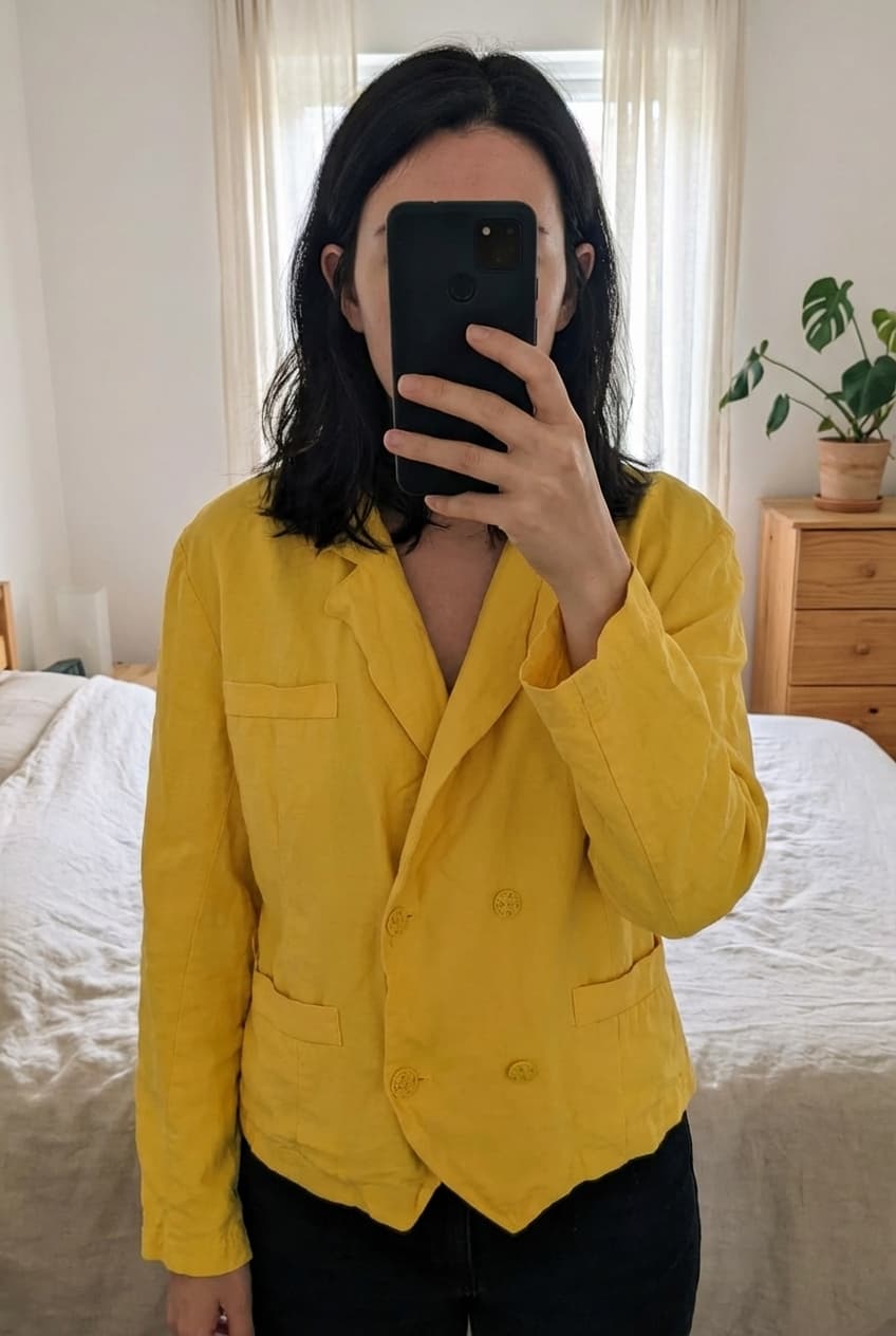 Veste vintage jaune debout, face à la caméra, dans une chambre à la lumière douce, avec une lumière naturelle filtrant par la fenêtre.