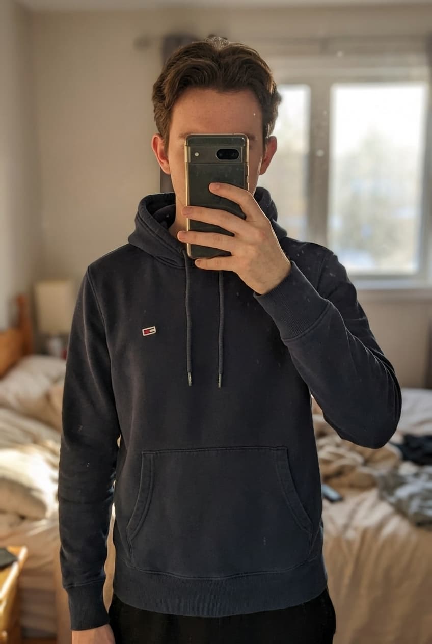 Sweat ommy Hilfiger debout, face à la caméra, dans une chambre à la lumière douce, avec une lumière naturelle filtrant par la fenêtre.