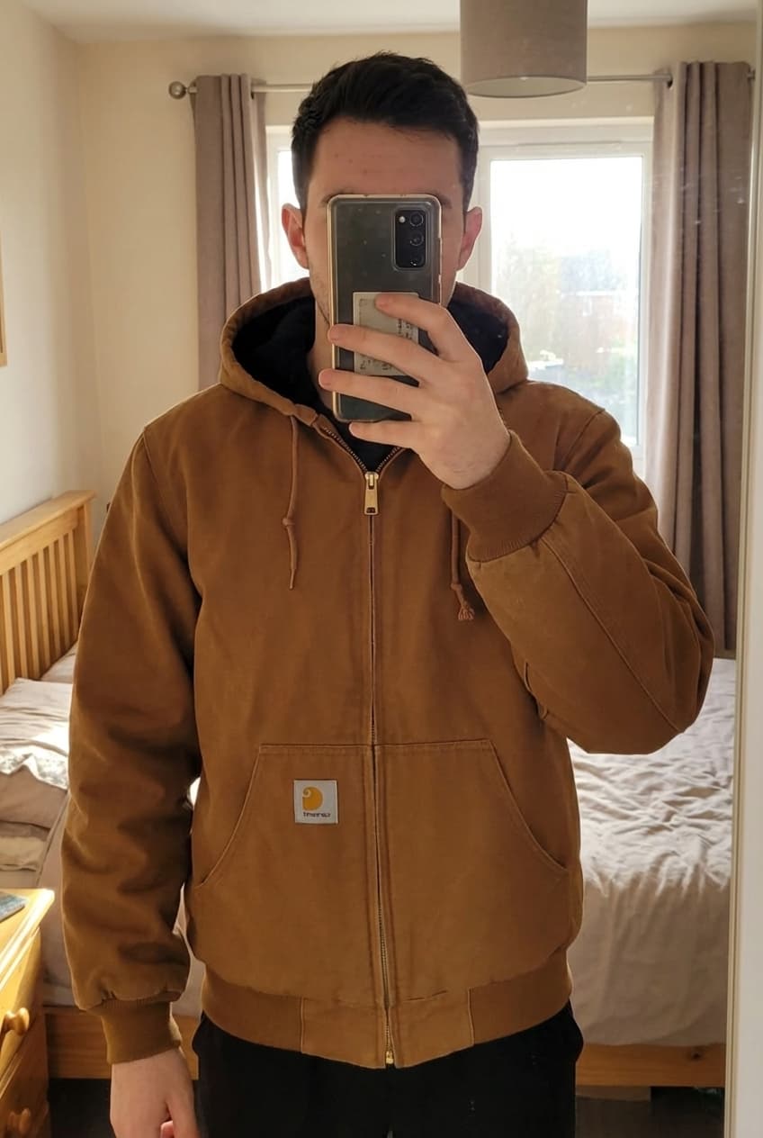 Veste Carhartt - M debout, face à la caméra, dans une chambre à la lumière douce, avec une lumière naturelle filtrant par la fenêtre.