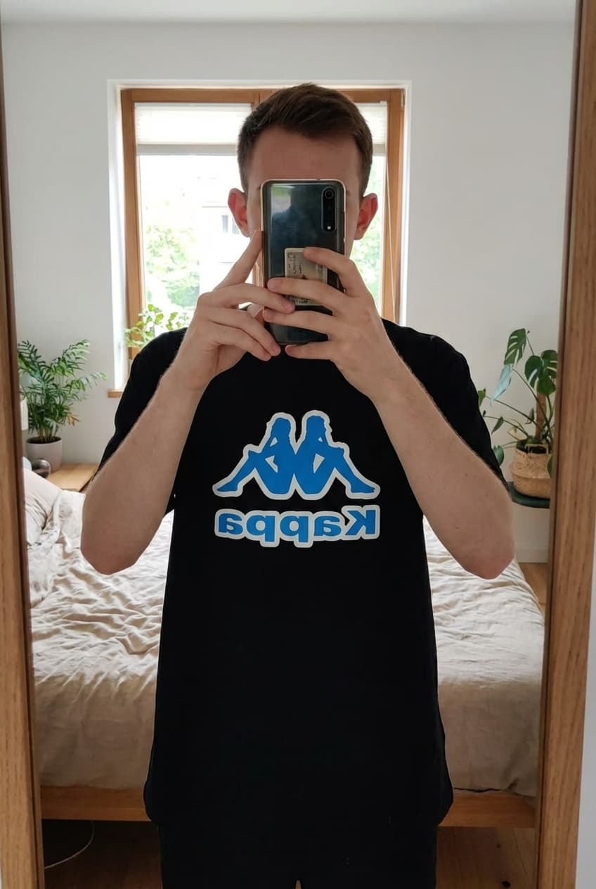 Une personne en T-shirt manches courtes Taille XXL debout, face à la caméra, dans une chambre à la lumière douce, avec une lumière naturelle filtrant par la fenêtre.