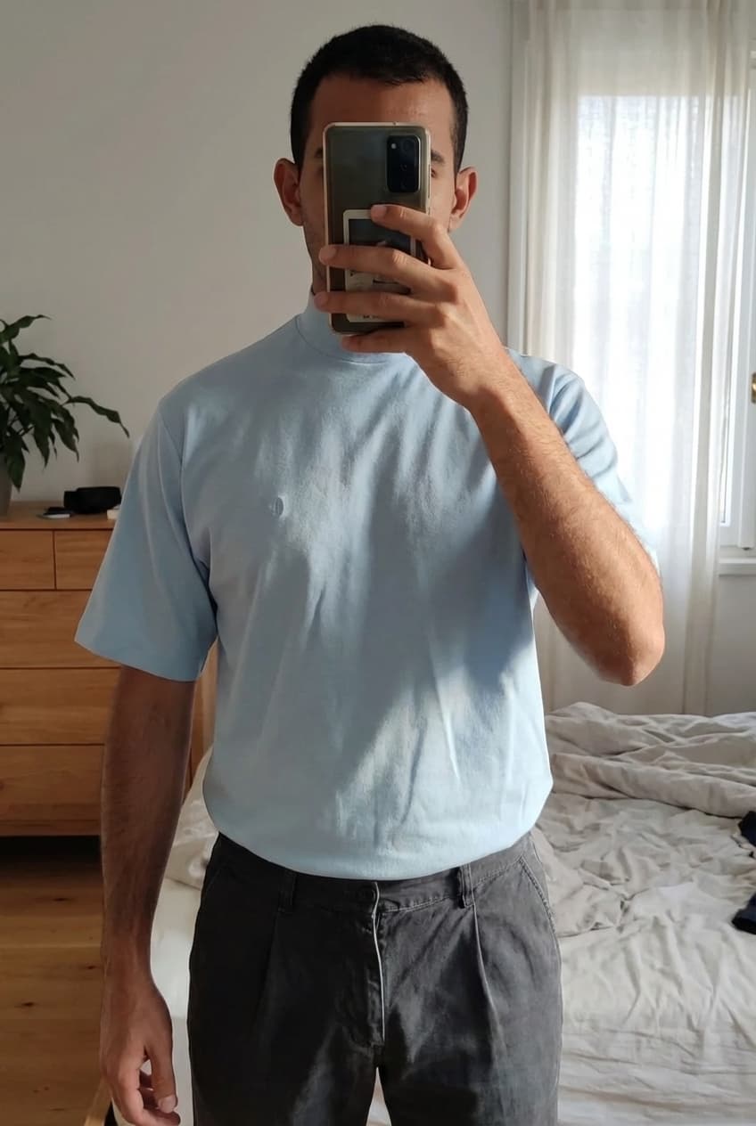 Une personne en T-shirt, col roulé, manche courte, conte of florence, taille 54 debout, face à la caméra, dans une chambre à la lumière douce, avec une lumière naturelle filtrant par la fenêtre.